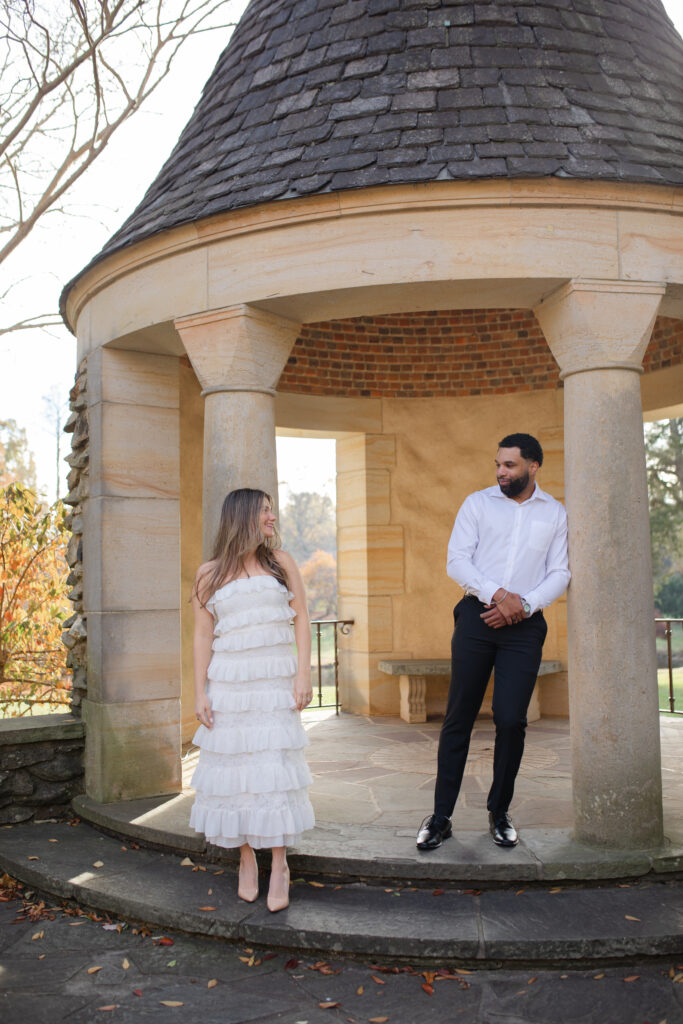 Man and woman stand a foot apart from one another under the rotunda at Graylyn Estate smiling at each other.