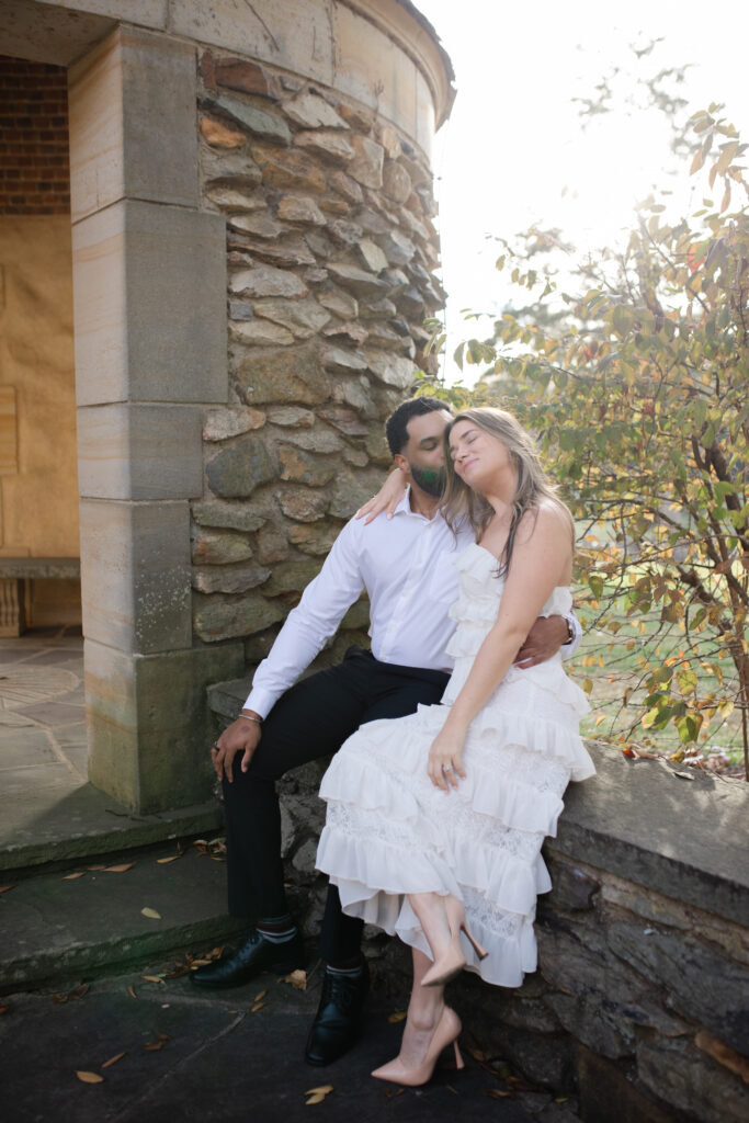 Man and woman sit together on wall cuddled together in the garden at Graylyn Estate. The man kisses the woman on the head.