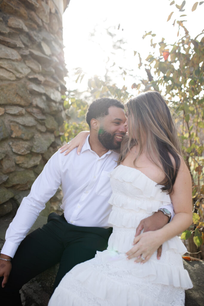 Close up of the man and woman as they look at one another smiling. They are cuddling as they sit on the wall in the garden.