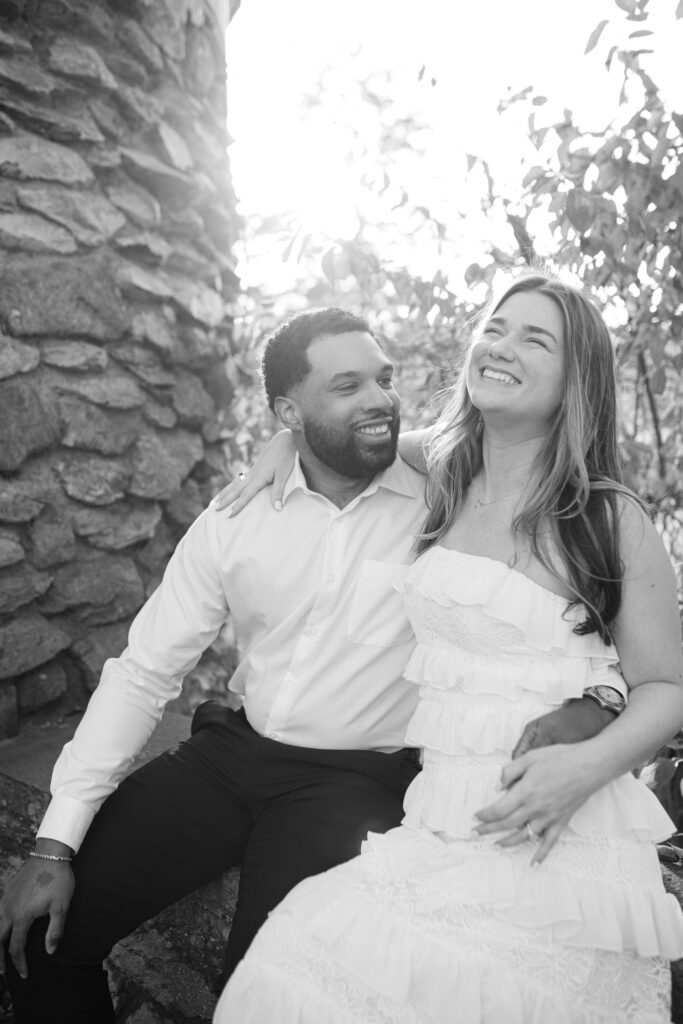 The woman looks away from the man smiling as they are cuddling together on the wall in the garden at Graylyn Estate.