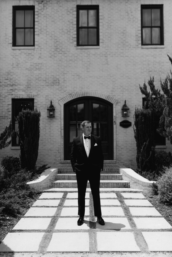 The groom stands outside the front door of The Bradford. His hands are in his pockets as he wears a black suit with a bow tie.