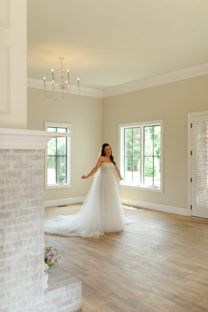 The bride smiles, holding out her hands excitedly, as she shows her bridesmaids her long, white, a-line wedding dress for the first time.