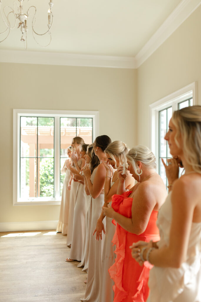 The bridesmaids, wearing various champagne colored dresses, look at the bride. Some smile and some cry happy tears.