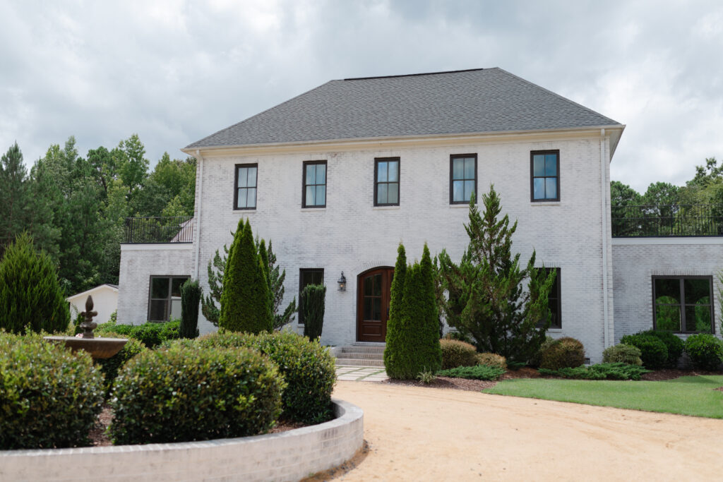 The outside of The Bradford in New Hill NC. Grey clouds are in the sky above the venue. The house is surrounded by lush greenery with a winding dirt road leading to the front door.