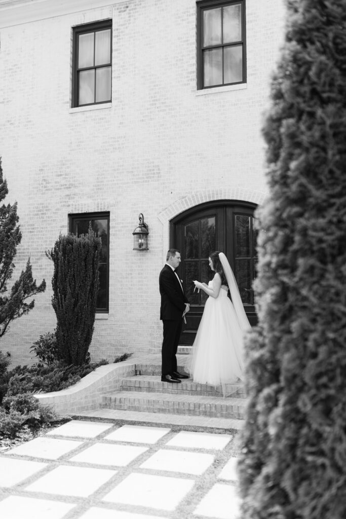 The bride reads her vows to the groom on the steps outside The Bradford in New Hill NC. The groom listens to her with a serious expression, holding back tears.