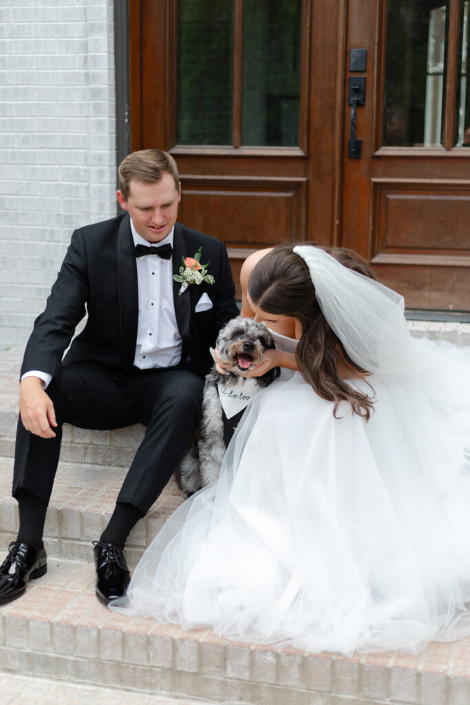 The bride and groom smile as they sit on the steps in front of The Bradford, petting and playing with their puppy.