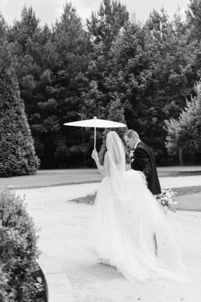 The groom helps hold the bride's dress and flowers as they walk outside the front of The Bradford. The bride holds a white parasol as they walk.