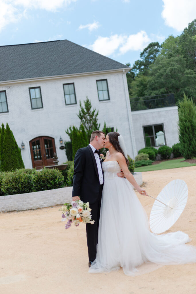 The bride and groom kiss in front of The Bradford in New Hill NC. The bride is holding her umbrella and the groom is holding her flowers. With their opposite arms they hold on to one another.