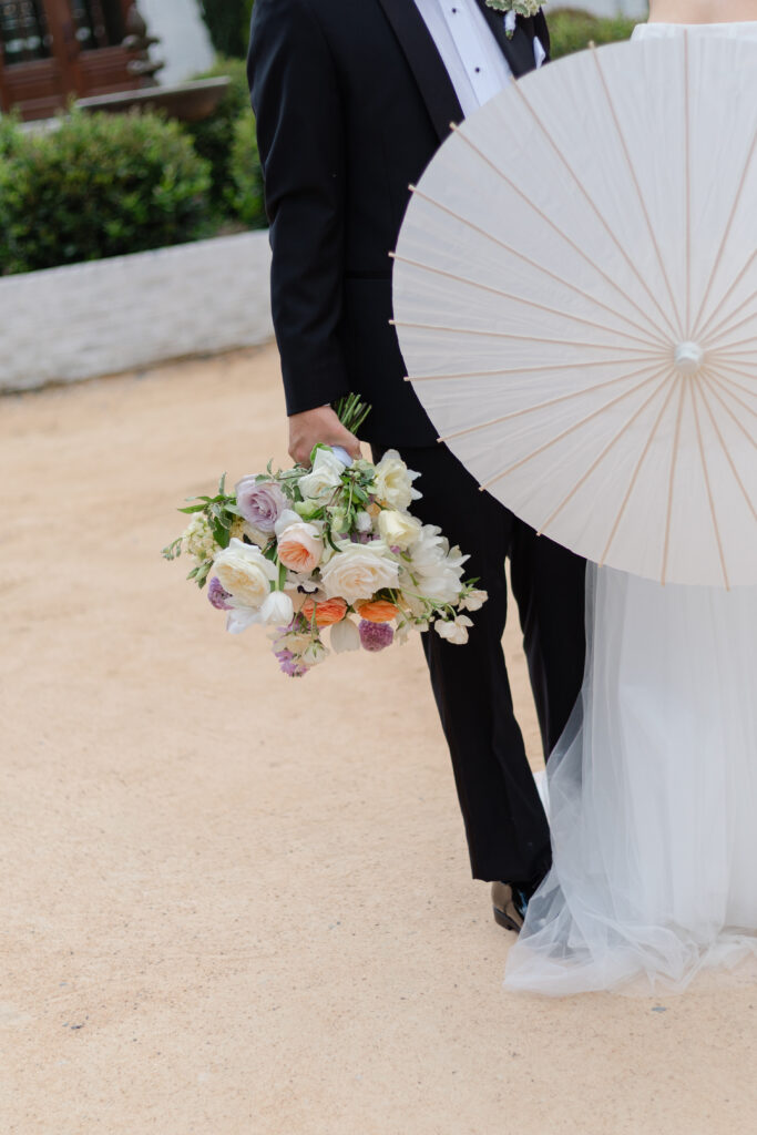 A close up of the bride's bouquet as the groom holds it. The flowers are cream, yellow, pink, orange, and purple.