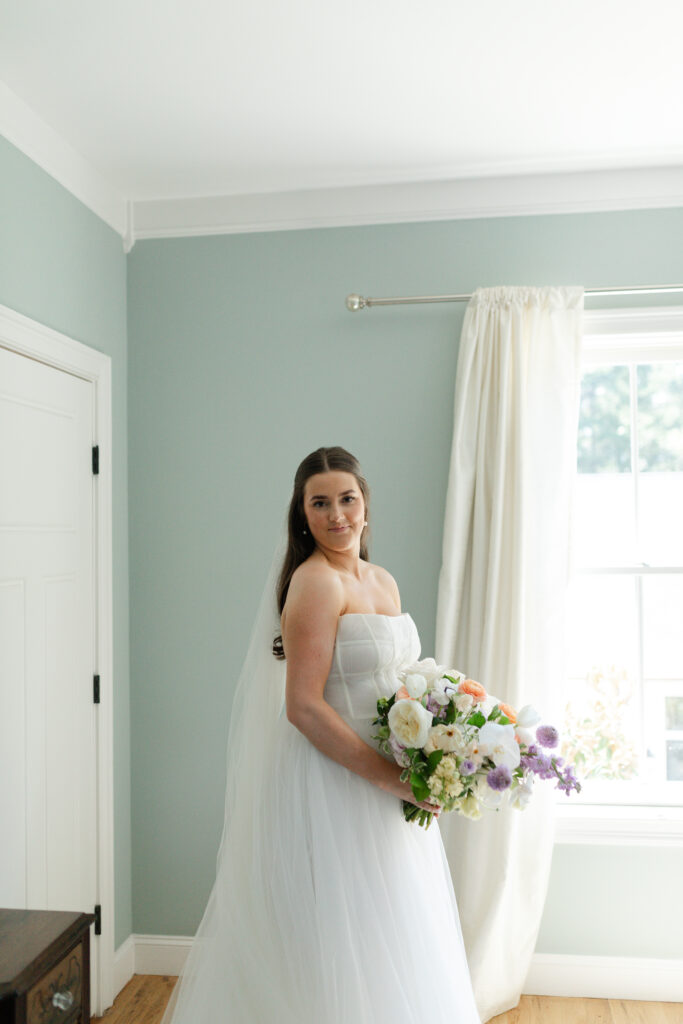 The bride looks at the camera, giving a side smile as she holds her bouquet in her bridal suite.