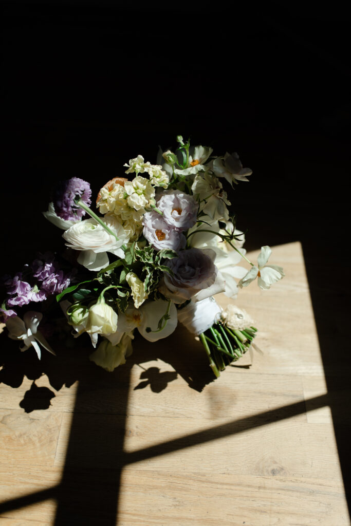 The bride's bouquet, containing purple, cream, yellow, and orange flowers, lies on the floor as light from the outside window pours over it.