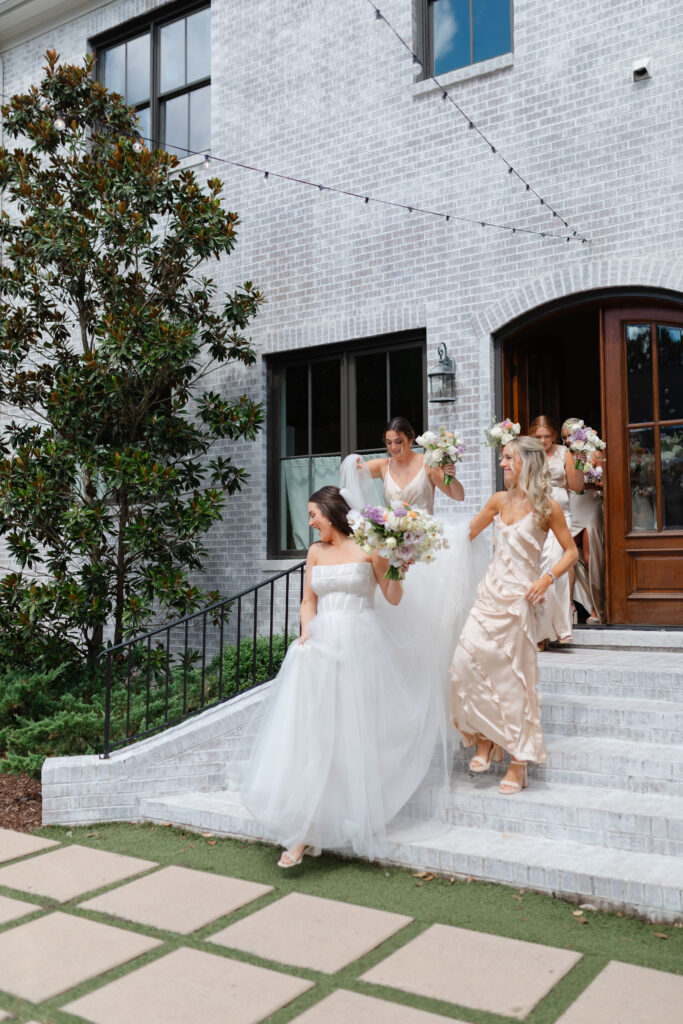 The bride walks down a flight of stairs outside. Two of her bridesmaids help carry her large dress. The other bridesmaids follow behind.