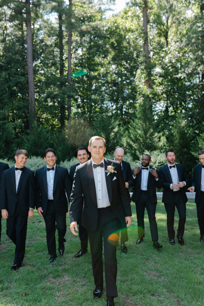 The groom walks through a green yard with his groomsmen, smiling at the camera as his friends walk and laugh behind him.