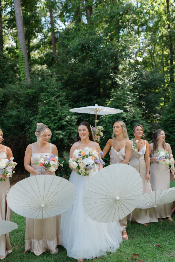 The bride smiles and laughs off to her right as her bridesmaids flock around her, laughing and smiling. They are all holding their bouquets and white parasols.