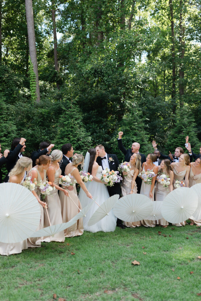 The bride and groom kiss as they are surrounded by their bridesmaids and groomsmen who look at them and cheer as they kiss.