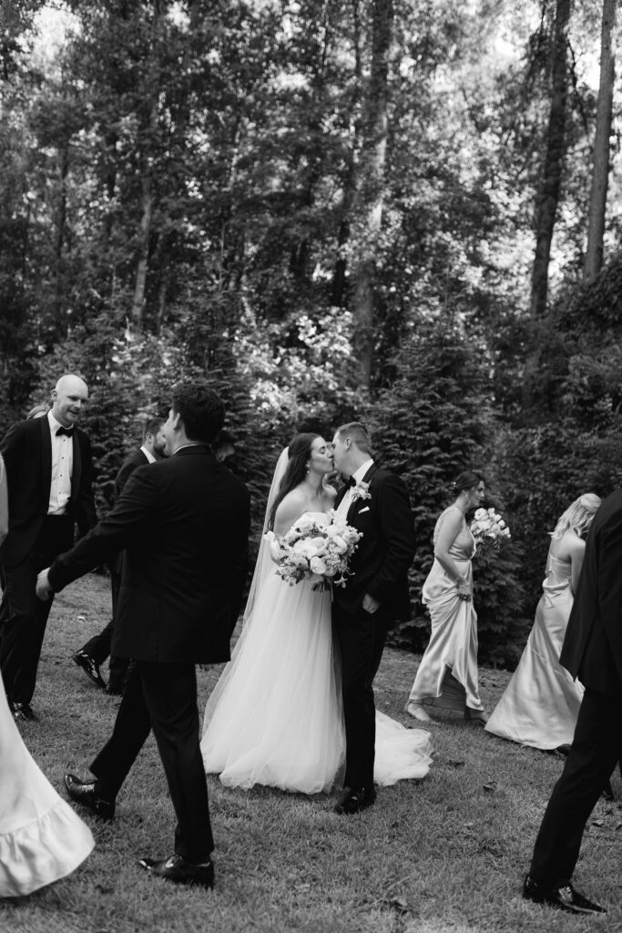 The bride and groom kiss as their bridesmaids and groomsmen swirl around them, smiling and laughing.