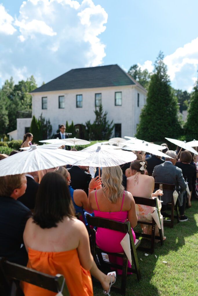 Guests are seated at the ceremony with white parasols shading them from the sun. In the background you can see The Bradford in New Hill NC.