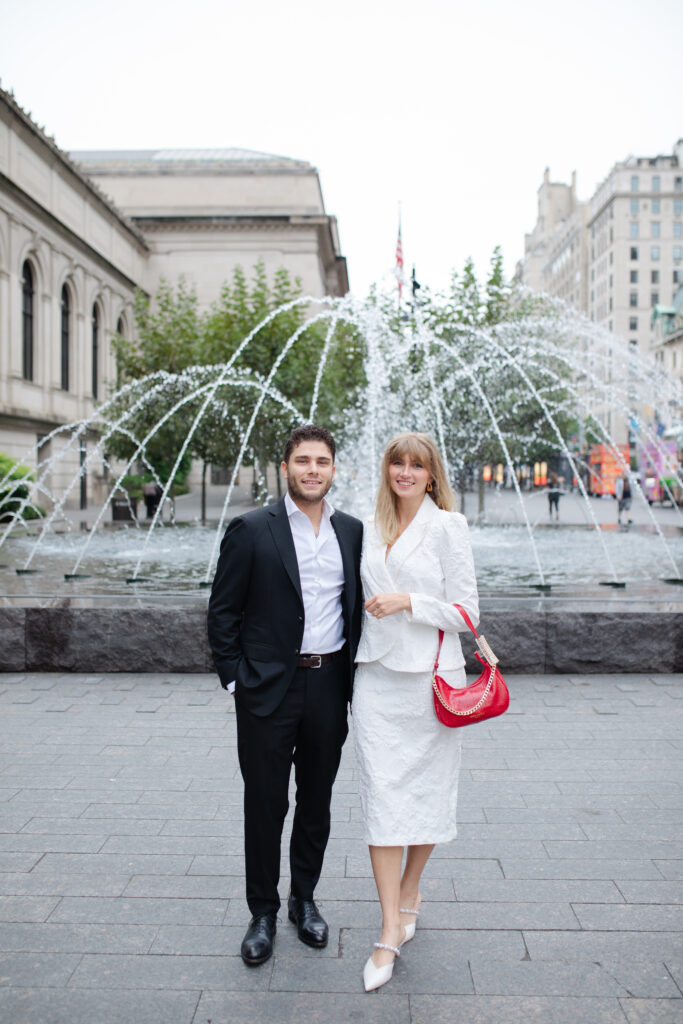 Man and woman stand side by side and arm in arm smiling at the camera. They are outside The MET for their engagement session.