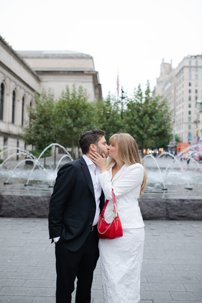 A man in a black suit kisses a woman in a white blazer and pencil skirt. She has a red bag. Behind them is a beautiful fountain. They are outside The MET.