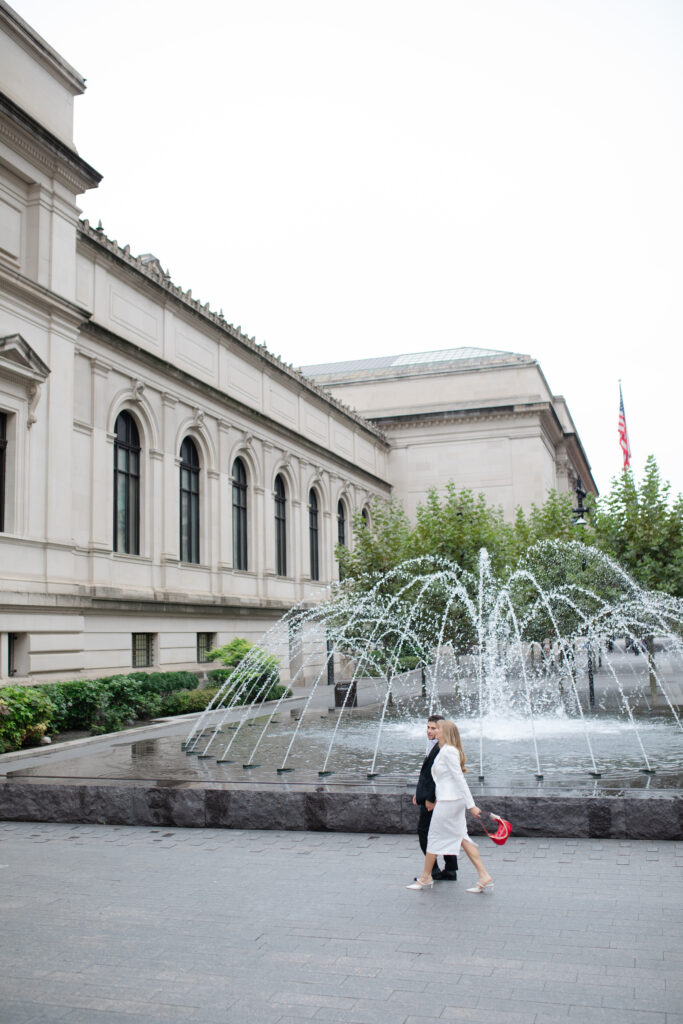 A man and woman walk arm and arm together outside the MET. Behind them is a gorgeous water fountain.