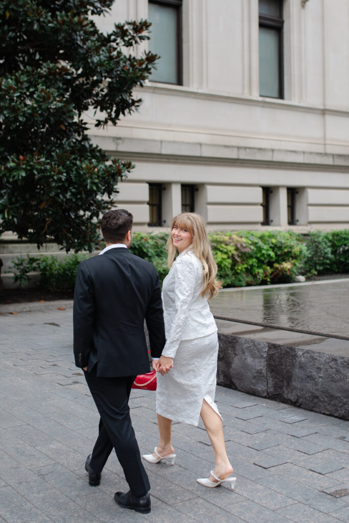 A man and woman walk hand in hand together outside The MET for their engagement session. The woman smiles over her shoulder at the camera as she walks.