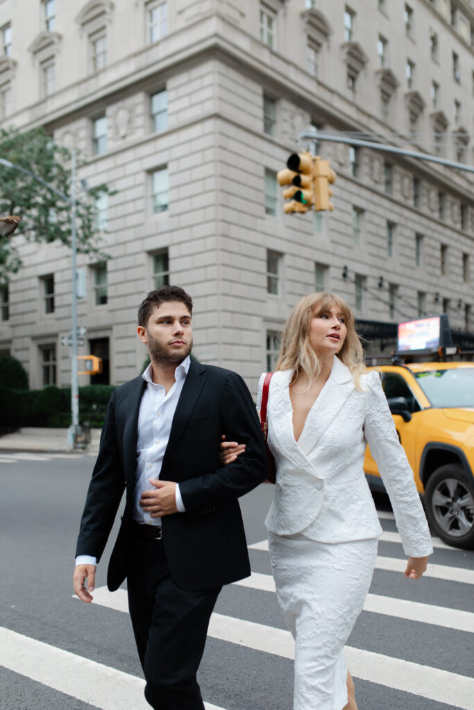 The man and woman walk across the street to The MET arm in arm. A yellow NYC taxi drives past them in the background.