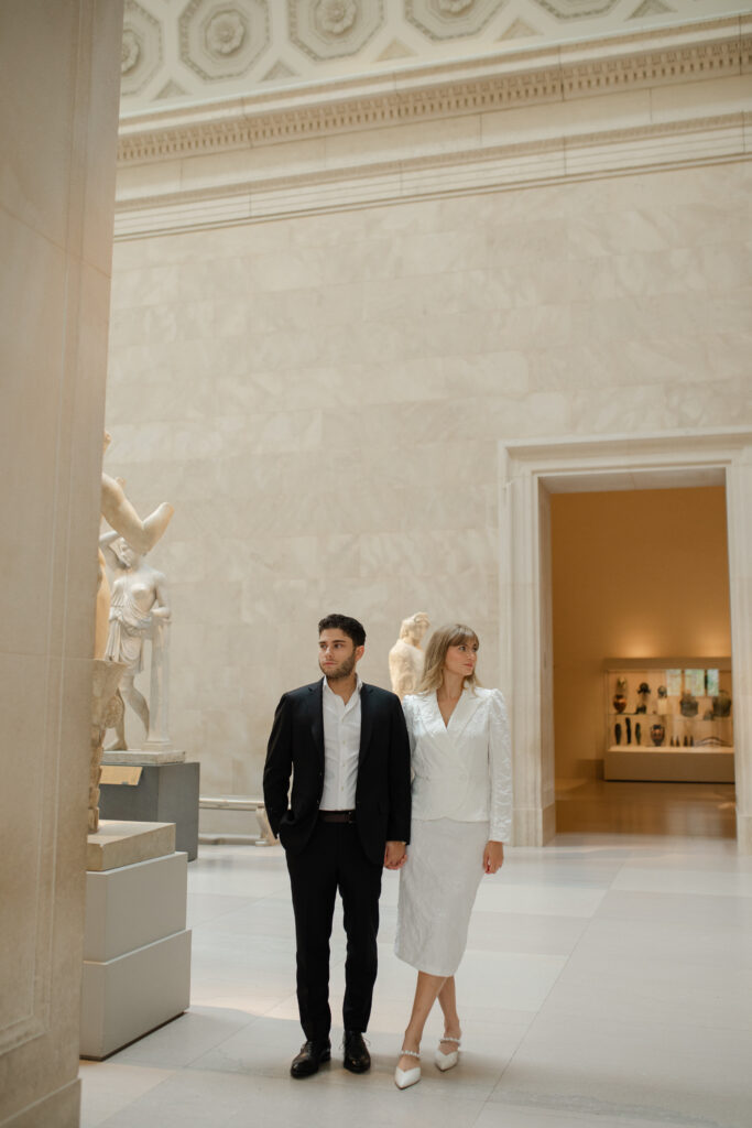 Man and woman inside the museum for their engagement session at The MET. They hold hands and look in opposite directions from one another, serious expressions on their face.