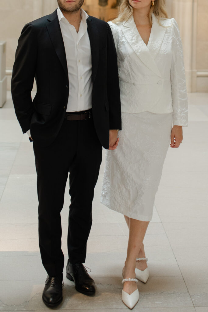 Close up photo of the man and woman's outfits. She is wear white heels with pearls on them, a white pencil skirt, and a white blazer with gold earrings. He is wearing a black suite with a white under shirt, no tie.