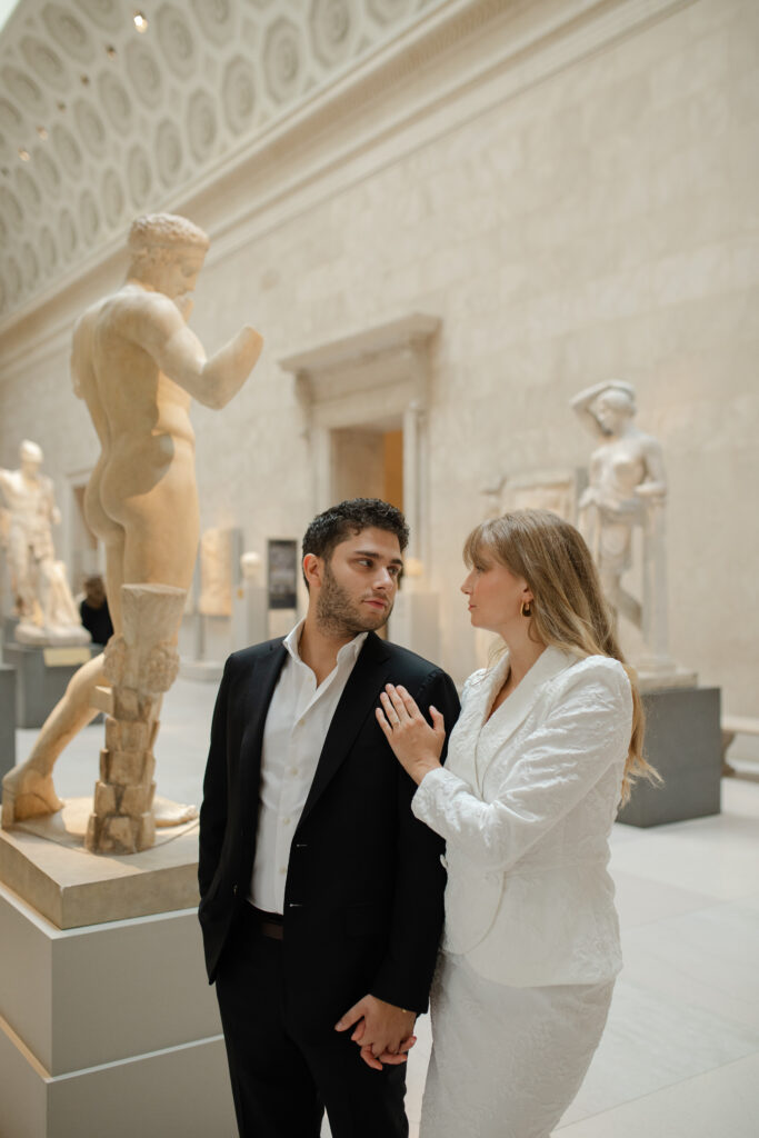 The man and woman stand side by side and hold hands together. She places her hand on his shoulder and they look lovingly into each others eyes. They are surrounded by beautiful white statues.