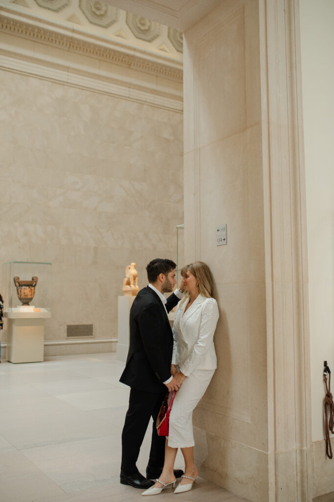 The woman leans against a wall in one of the grand corridors within The MET. He stands in front of her with his hand on the wall, enclosing her. Their faces are very close as they smile at one another.