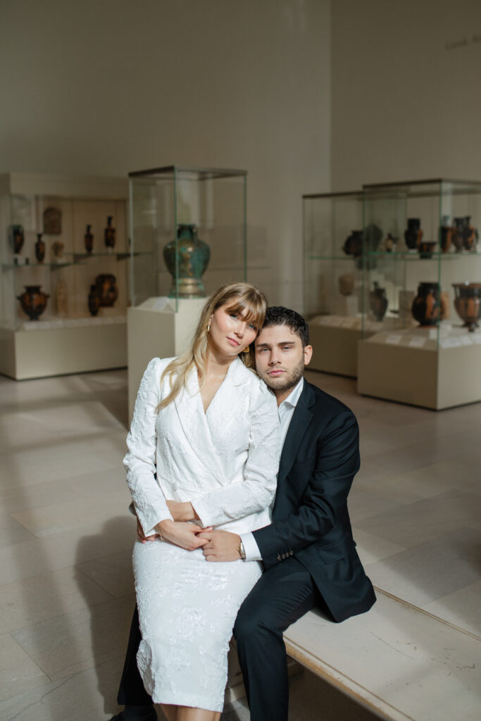 The man and woman are sitting on a bench in a hall filled with beautiful vases. She sits on his lap and leans her head against his. They both look at the camera with a serious expression.