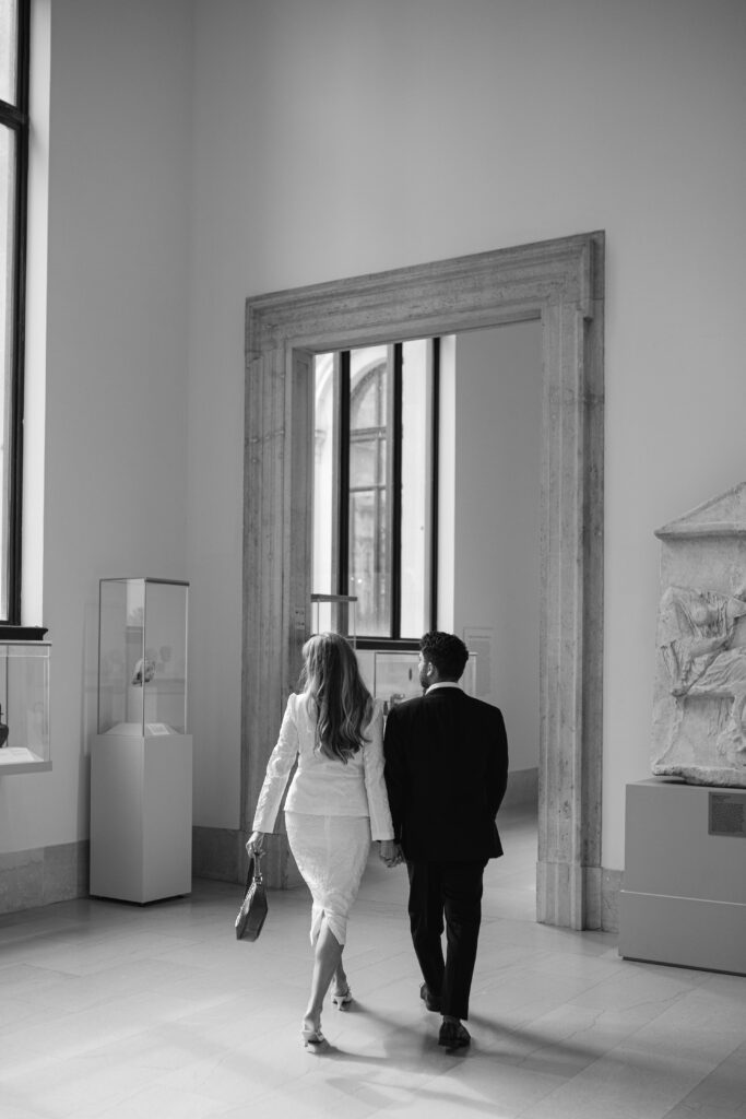 The man and woman walk away hand in hand, looking off to their left as they pass through the gallery.