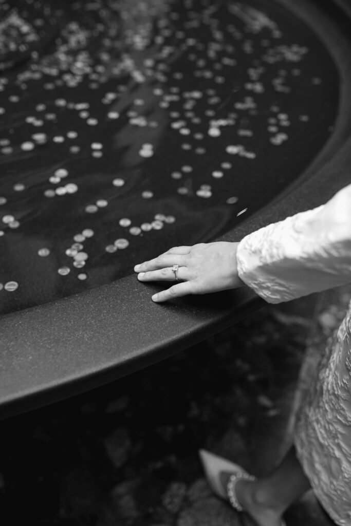 A detail shot of the woman's hand, showing off her engagement ring. She places her hand on the edge of a water fountain. There are many pennies inside the fountain.